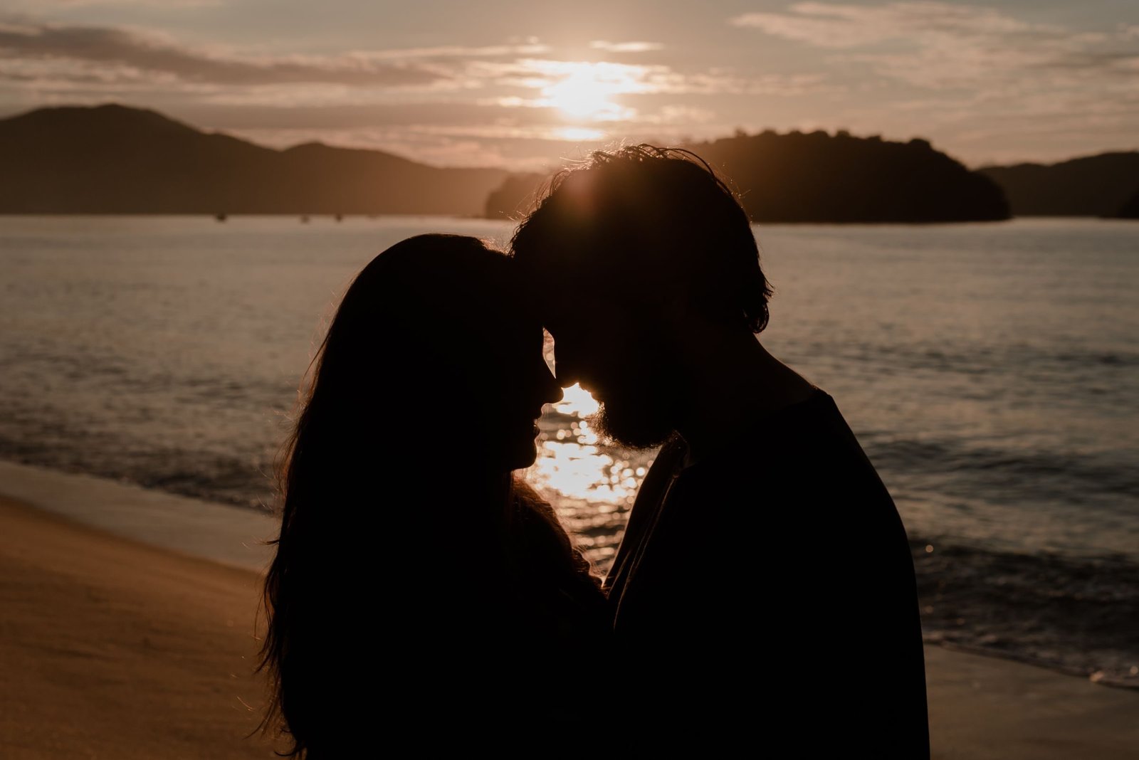 A couple in silhouette, foreheads touching, against the sea and the setting sun